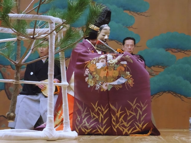 Intérprete de Noh en la obra Matsukaze, detrás de un pequeño pino y llevando un kimono violeta y una máscara noh