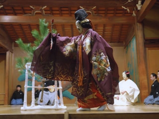 intérprete de Noh de cuerpo entero, mirando hacia la isquierda, en la obra Matsukaze, llevando un kimono violeta, máscara noh y un abanico