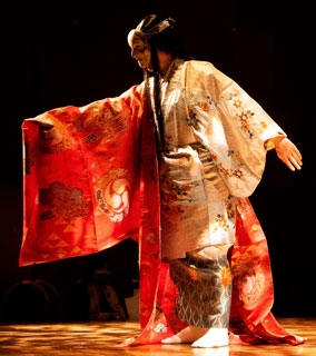 Kanji Shimizu interpretando noh con una máscara noh, el brazo delerecho elevado y un kimono naranja y blanco