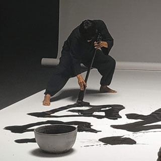 Mitsuru Nagata con un pincel de grande dimensiones escribiendo shodo en un gran papel en el suelo