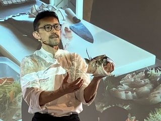 Uno de los ponentes enseña partes de un pescado bajo una proyección de una imagen de una tabla de cortar