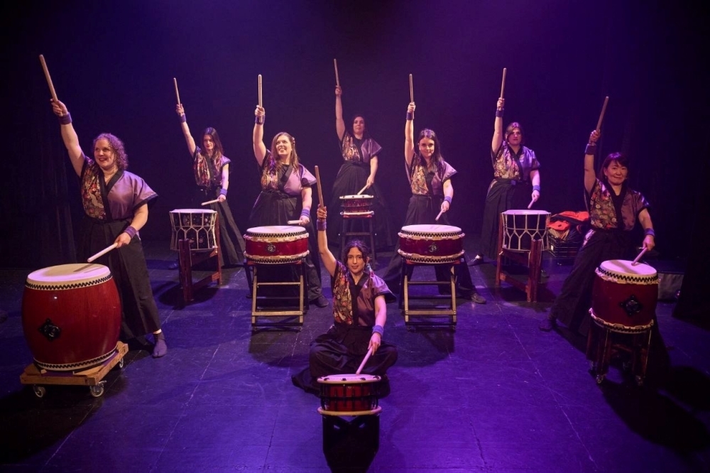 Las 8 miembros de Murasaki taiko ante sus tambores con las baquetas bachi en alto en una actuación