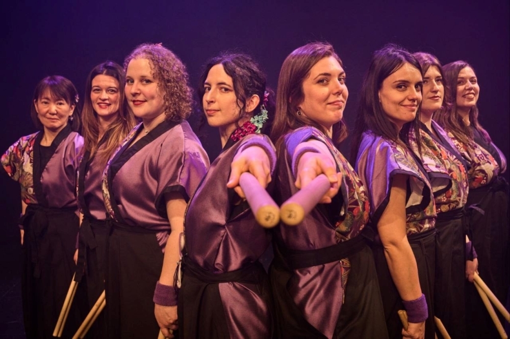 Las 8 miembros de Murasaki Taiko colocadas en triángulo y dos de ellas enseñan sus baquetas bachi