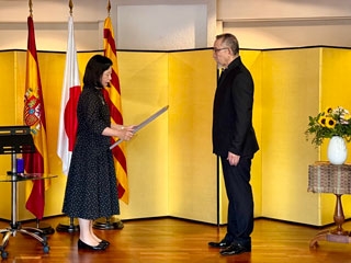 La Cónsul General lee el diploma de la condecoración al Sr. Sotoo ante tres banderas y un biombo dorado y junto a un jarrón de flores